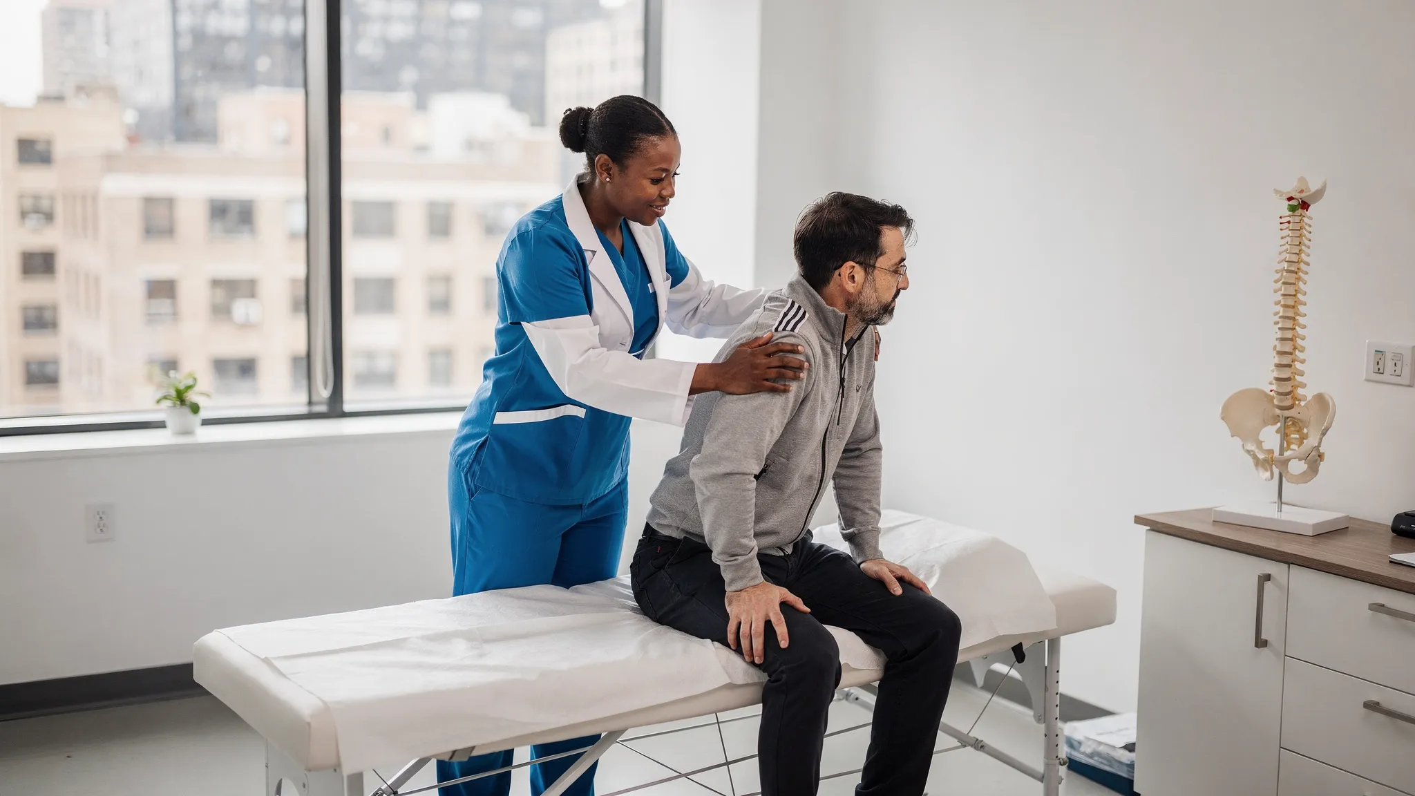 A clinician in a modern Manhattan clinic exam room demonstrates a simple hip mobility test to a patient while pointing to a spine and pelvis model on a nearby counter. The setting looks clean and professional, with a treatment table, neutral decor, and no visible branding.