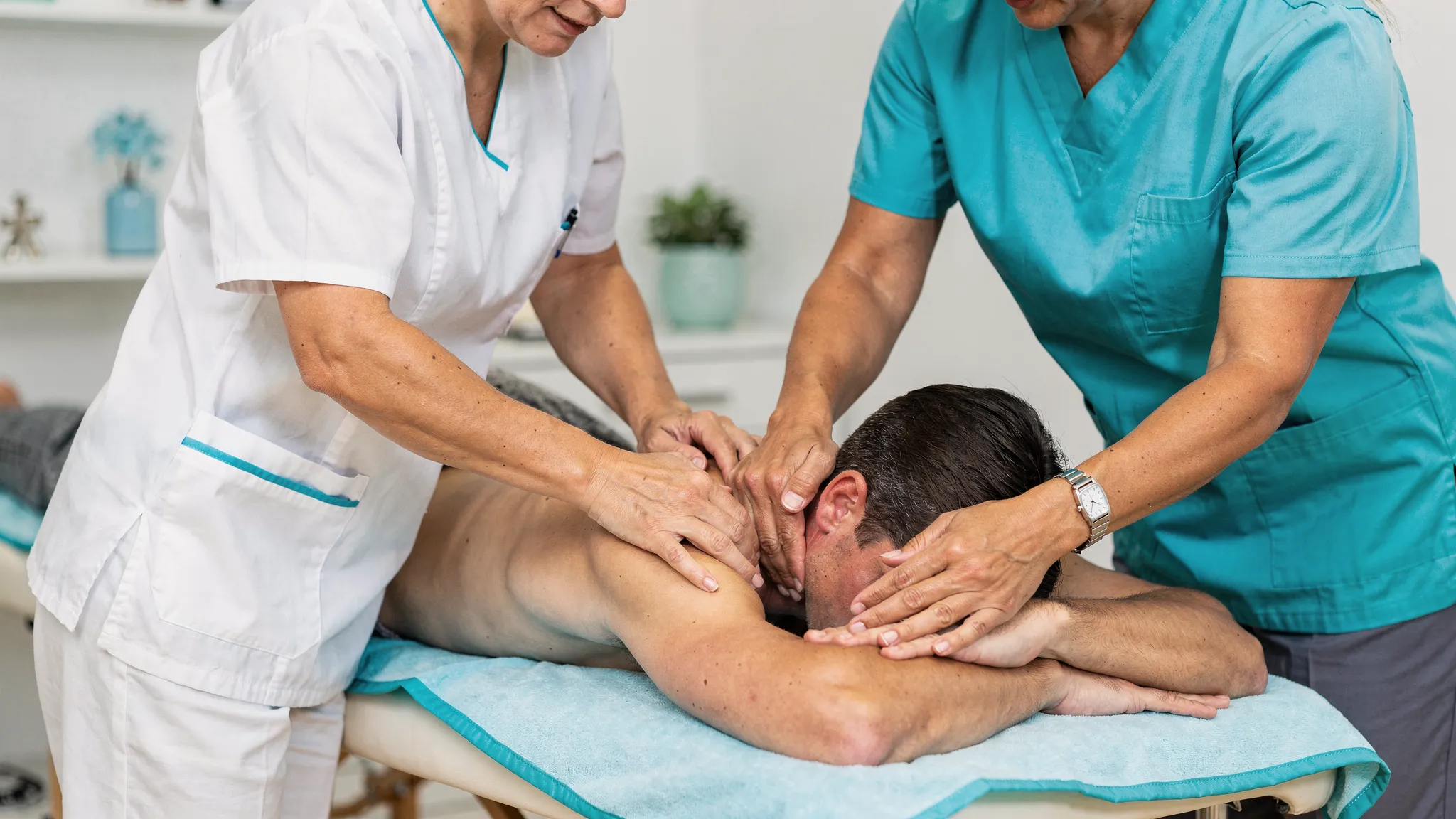 A clinician palpates a patient’s upper back and shoulder area on a treatment table while another provider demonstrates gentle soft tissue work on the neck and trapezius, illustrating coordinated chiropractic and massage-style care in a calm clinic room.