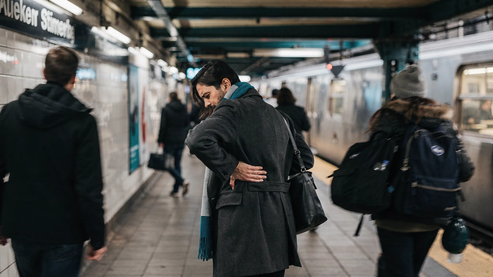 A New York City commuter standing on a subway platform with one hand on their lower back, looking uncomfortable, while other commuters walk by; the scene emphasizes everyday back pain in NYC and the need for trusted care.