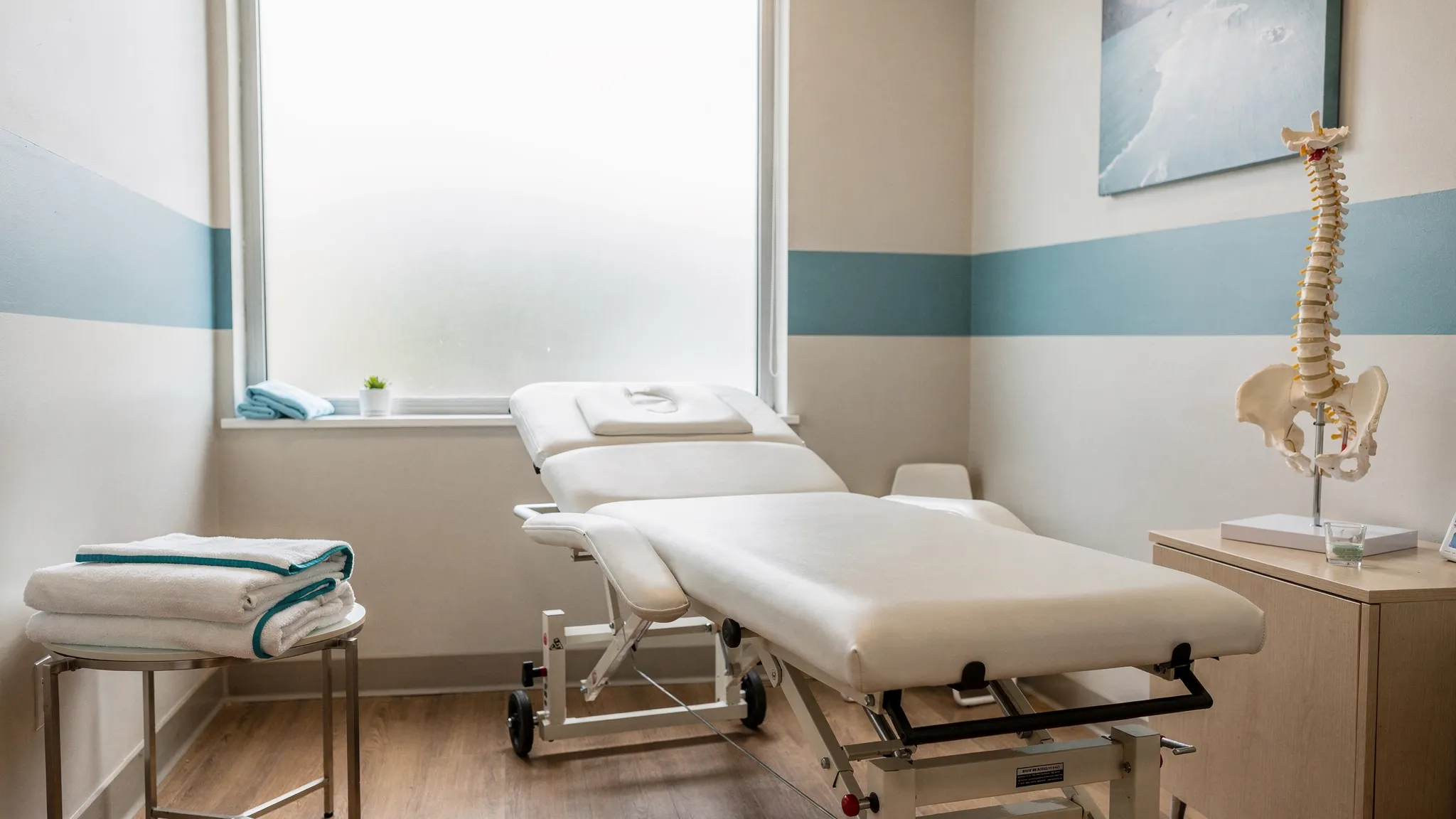 A calm chiropractic treatment room with an exam table, anatomical spine model, folded towels, and natural light, prepared for a weekend patient appointment.