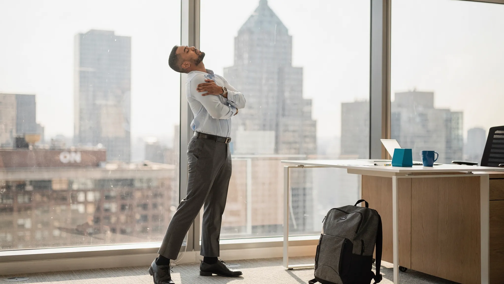 A Manhattan office worker standing near a desk while gently stretching the neck and shoulders, with a city skyline visible through the window and a backpack resting nearby.
