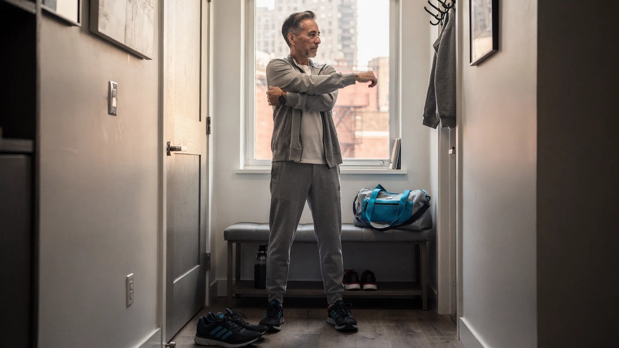 An adult New Yorker in comfortable work clothes does a gentle standing hip and shoulder mobility routine in a quiet apartment entryway before leaving for the day. A gym bag, walking shoes, and a city window view reinforce a realistic wellness routine that fits into a packed schedule.